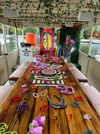 Party boat interior on a canal with a long wooden table covered in colorful leis, beads, wristbands and a '25' birthday cake, ivy-draped ceiling, a 'Survivor' banner and waterfront views.