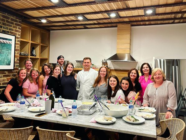 Smiling group and a chef gathered around a marble island in a modern farmhouse kitchen with wood‑paneled ceiling, exposed brick and stainless range hood — private cooking class/dinner party.