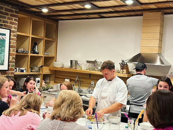 Chef leading a hands-on cooking class at a marble-topped island, small group of adults sampling dishes in a cozy open kitchen with wood shelving, exposed brick and stainless range hood