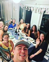 Selfie of a smiling group of friends around a set table at a home dinner party, wine bottles and plates on a blue tablecloth with a floral celebration banner overhead.