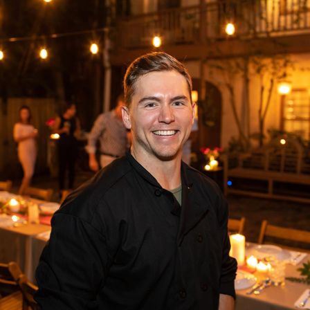 Smiling chef in a black jacket at a cozy outdoor patio dinner party with string lights and candlelit tables