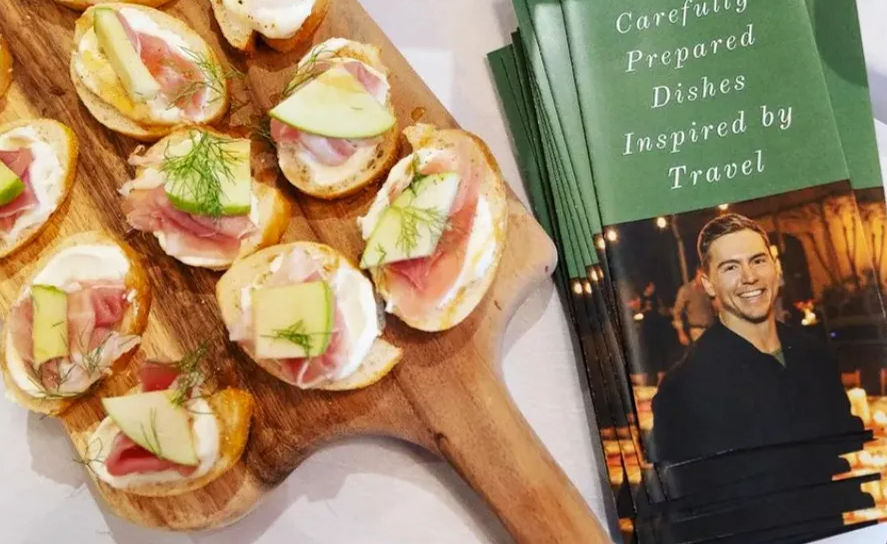 Wooden serving board of bite-size crostini topped with cream cheese, prosciutto, thin apple slices and dill, set next to a stack of green travel-inspired recipe booklets featuring a smiling chef photo.