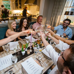 Cheerful friends making a white-wine toast at a home dinner party in a modern kitchen dining room, table set with printed menus, candles and small floral centerpieces.