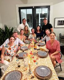 Cheerful group of friends gathered for a home dining room dinner party around a table with patterned plates, woven chargers, tulip centerpiece, candles and wine glasses, black French doors in the background.