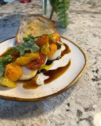 Caprese-style appetizer: fresh mozzarella rounds topped with roasted cherry tomatoes, fresh basil, balsamic glaze and olive oil, served with toasted sourdough on a marble countertop.