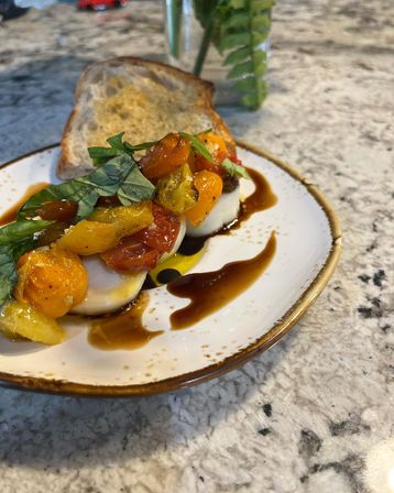 Caprese-style appetizer: fresh mozzarella rounds topped with roasted cherry tomatoes, fresh basil, balsamic glaze and olive oil, served with toasted sourdough on a marble countertop.
