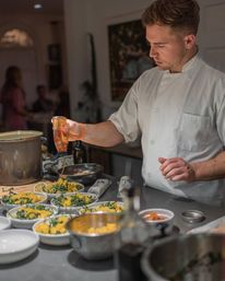 Professional chef in a white coat pouring dressing over multiple salad bowls of greens and yellow vegetables on a stainless prep counter in a busy restaurant kitchen.