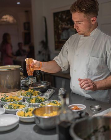 Professional chef in a white coat pouring dressing over multiple salad bowls of greens and yellow vegetables on a stainless prep counter in a busy restaurant kitchen.