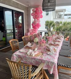 Coastal covered patio dining table styled for a pink-themed celebration with a tall pink balloon garland, floral pink tablecloth, pastel place settings, fresh floral centerpieces and a beach/ocean view