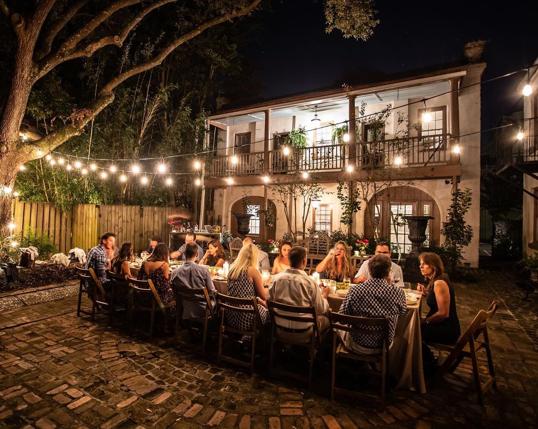 Al fresco evening dinner on a cobblestone courtyard under string lights, a long table of guests dining in front of a two-story house with a balcony and warm cozy lighting.