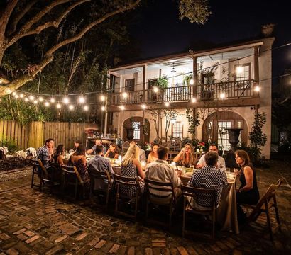 Al fresco evening dinner on a cobblestone courtyard under string lights, a long table of guests dining in front of a two-story house with a balcony and warm cozy lighting.