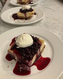 Blueberry cheesecake slice topped with glossy blueberry compote and a dollop of whipped cream on a white plate, red berry sauce pooling on a marble tabletop with more plated desserts in the background.