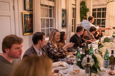 Lively dinner party in a cozy home dining room — guests seated along a long table with wine bottles, water, plates and white hydrangea centerpieces as a woman passes a serving bowl.