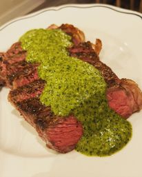 Close-up of juicy medium-rare ribeye steak slices topped with vibrant green chimichurri sauce on a white plate.