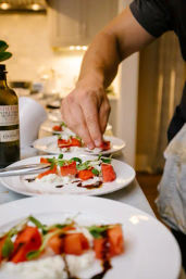 Hand plating vibrant watermelon and burrata salad with balsamic glaze and microgreens on white plates at a kitchen counter, olive oil bottle visible.