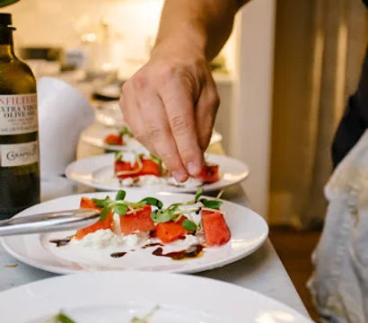 Hand plating vibrant watermelon and burrata salad with balsamic glaze and microgreens on white plates at a kitchen counter, olive oil bottle visible.