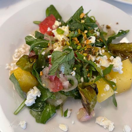Mediterranean-style summer arugula salad with watermelon and yellow melon cubes, crumbled feta, chopped pistachios, roasted peppers and pickled shallots on a white plate.