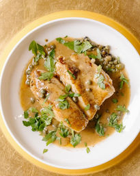 Top-down view of seared chicken breasts in a creamy lemon-caper sauce, sprinkled with fresh parsley on a white plate