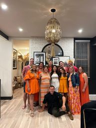 Smiling group of friends in orange and white outfits posing for an indoor celebration in a modern living room with a rattan pendant light, white brick accent wall, framed art and light wood floors.