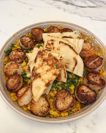 Ceramic bowl of pan-seared scallops arranged around creamy charred corn and chopped cilantro, topped with folded grilled flatbread on a marble countertop