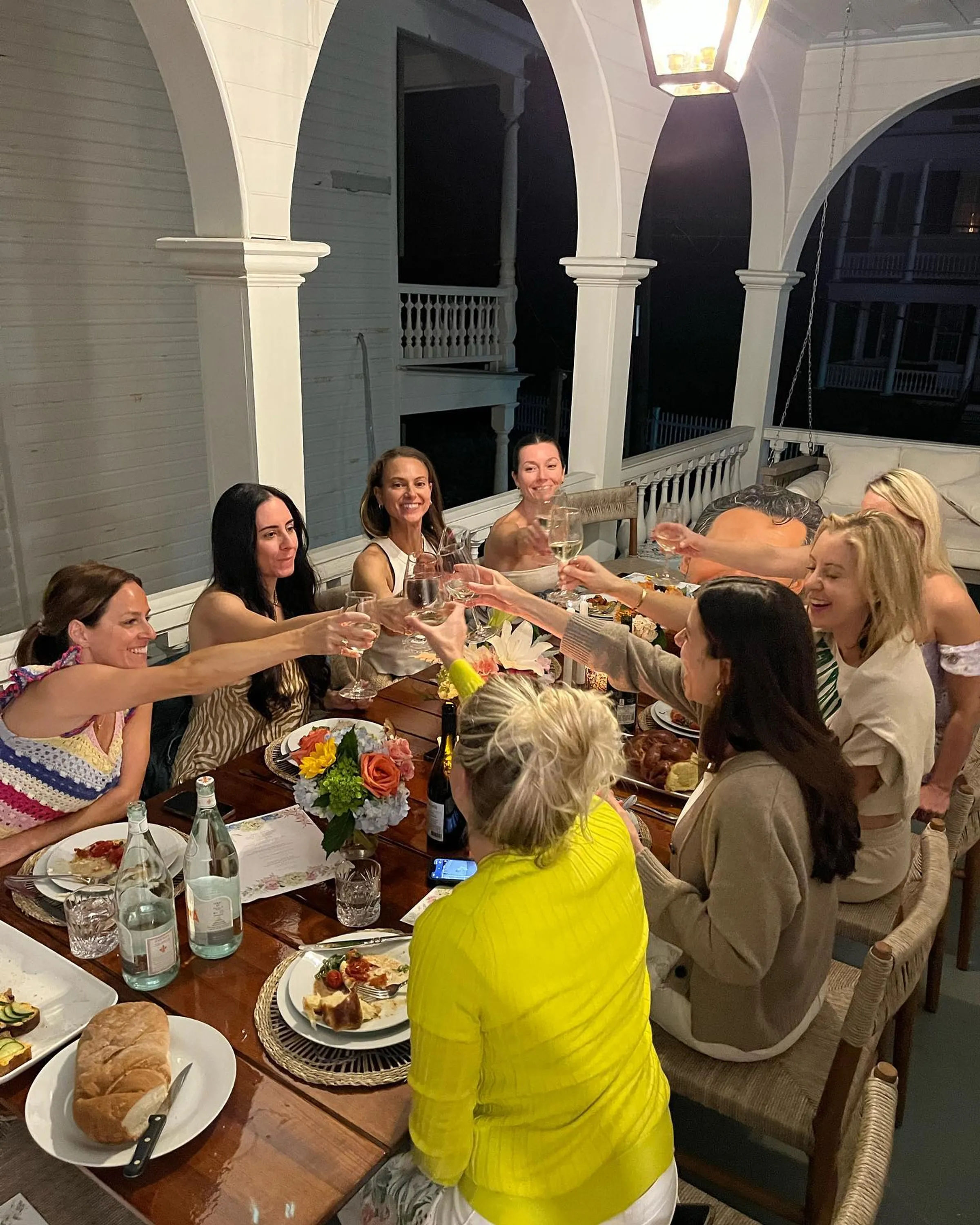 Lantern-lit outdoor dinner party on a white arched porch — a group of women toasting with wine around a long wooden table set with plates, bread and a floral centerpiece.