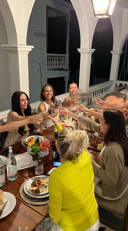 Lantern-lit outdoor dinner party on a white arched porch — a group of women toasting with wine around a long wooden table set with plates, bread and a floral centerpiece.