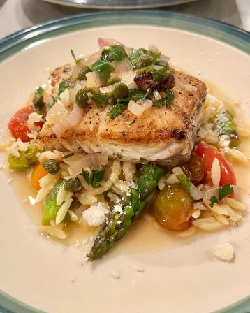 Golden-seared white fish fillet atop orzo with asparagus, blistered cherry tomatoes, capers, chopped herbs and grated cheese in a light lemon-butter sauce, Mediterranean-inspired.