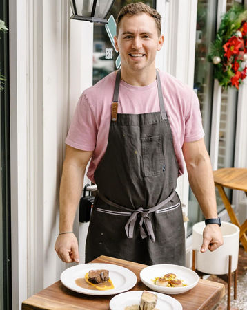 Smiling chef in a pink t-shirt and gray apron presenting three plated gourmet dishes on a wooden outdoor table outside a bistro decorated with a holiday wreath