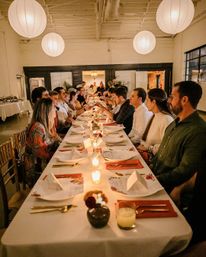 Candlelit communal dinner party in a loft-style event space with round paper lanterns overhead, a long table set with menus, gold flatware and guests seated facing each other.
