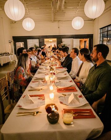 Candlelit communal dinner party in a loft-style event space with round paper lanterns overhead, a long table set with menus, gold flatware and guests seated facing each other.