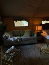 Cozy glamping tent interior with a gray sofa draped in a blanket, textured pillows, glowing bedside lamp beside a woven bed, rolled towels on a chair, potted plant and jute rug.