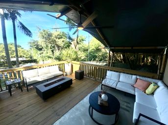 Sunny covered tropical wooden deck patio with white cushioned sectionals and colorful throw pillows, round coffee table, rectangular fire pit table, ceiling fan, potted plant, and palm trees with lush green foliage beyond the railing.