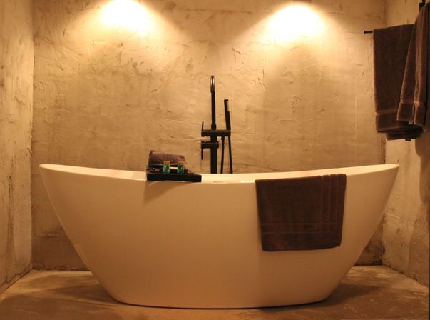 Warm-lit modern freestanding white bathtub with black floor-mounted faucet, textured concrete walls, dark brown towels on the tub and wall, and a bath caddy with toiletries.