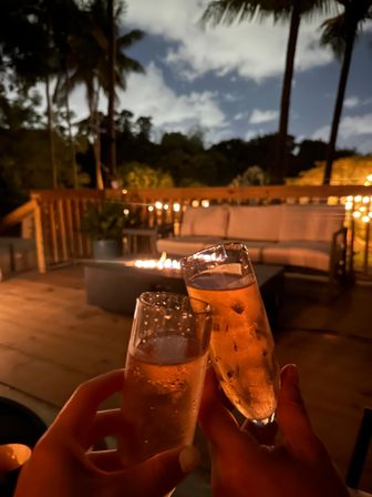 Two champagne flutes clinking on a cozy outdoor patio at night, fire pit and string lights on a palm‑fringed deck with cushioned sofa.