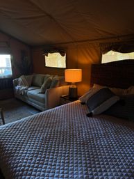 Cozy glamping tent interior with quilted bed and woven headboard, decorative pillows, bedside lamp casting a warm glow, and a soft sofa by canvas windows.