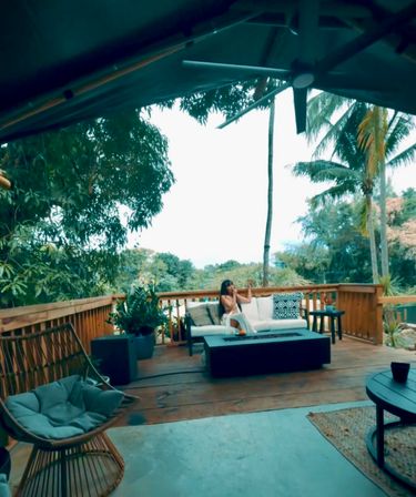 Tropical wooden deck lounge with palm trees and lush greenery; a person relaxes on a white outdoor sofa beside a coffee table and rattan hanging chair under a ceiling fan.