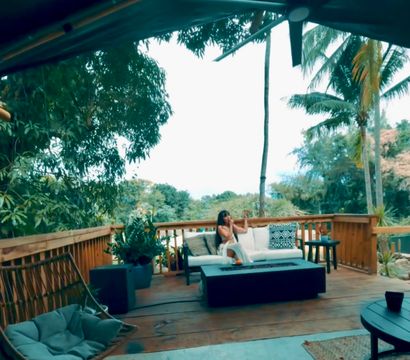 Tropical wooden deck lounge with palm trees and lush greenery; a person relaxes on a white outdoor sofa beside a coffee table and rattan hanging chair under a ceiling fan.