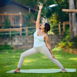 Person doing outdoor yoga on a green mat in a lush tropical backyard, wearing light activewear and holding a reverse warrior pose with one arm reaching upward and visible arm tattoos