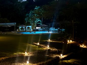 Blue-lit outdoor pool at night with cabanas and palm trees, a winding stone pathway edged by warm garden lights
