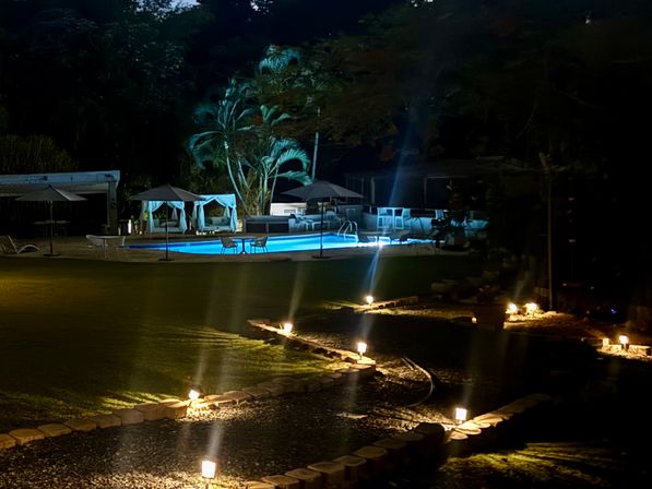 Blue-lit outdoor pool at night with cabanas and palm trees, a winding stone pathway edged by warm garden lights