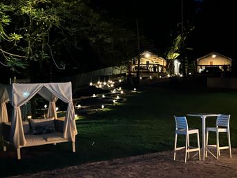 Nighttime tropical garden with a white draped cabana daybed, glowing pathway lights up stone steps to illuminated bungalows, and a small patio table with two high chairs — cozy outdoor lounge scene.