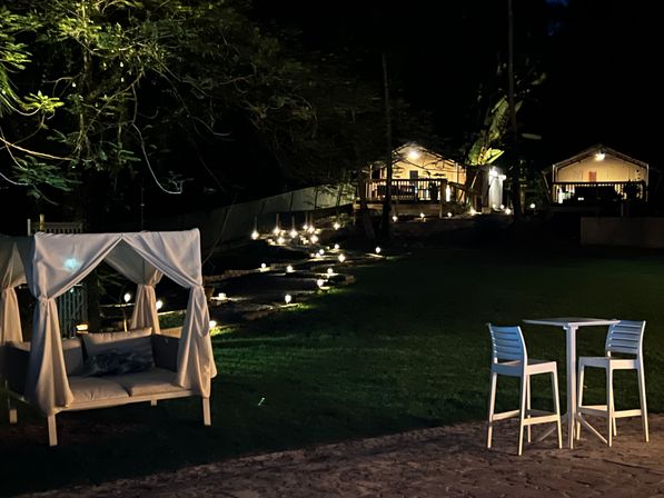 Nighttime tropical garden with a white draped cabana daybed, glowing pathway lights up stone steps to illuminated bungalows, and a small patio table with two high chairs — cozy outdoor lounge scene.