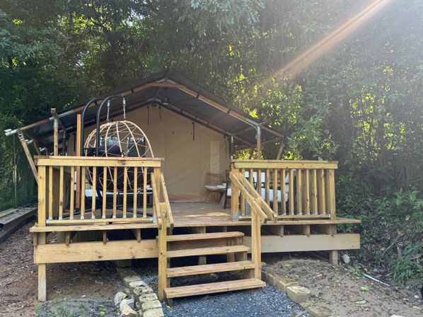 Sunlit glamping tent on a wooden deck in a leafy forest, featuring a hanging egg chair and wicker seating on the outdoor porch.