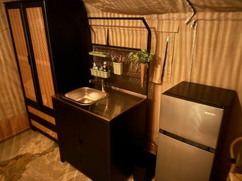 Compact kitchenette inside a canvas glamping tent with a stainless sink and countertop, black rattan-accented storage cabinet, hanging organizer with plants and toiletries, and a small silver refrigerator under warm lighting.