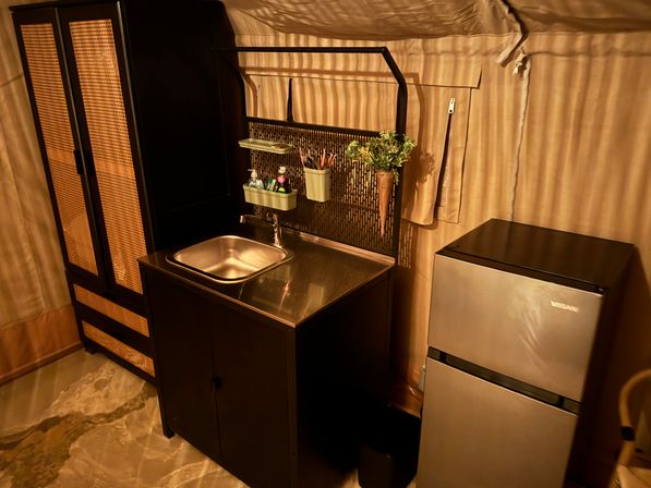 Compact kitchenette inside a canvas glamping tent with a stainless sink and countertop, black rattan-accented storage cabinet, hanging organizer with plants and toiletries, and a small silver refrigerator under warm lighting.