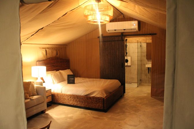 Cozy luxury glamping tent bedroom with woven rattan bed, bedside lamp and armchair, sliding barn door to en-suite bathroom, warm ambient lighting and concrete floor.