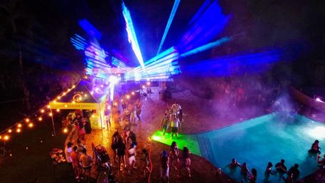 Nighttime outdoor pool party with people dancing on a wet patio under bright blue laser beams and warm string lights, a cocktail tent nearby and swimmers in a glowing illuminated pool