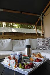 Cozy outdoor breakfast tray on a patio table: white tray with fresh grapes, plums and strawberries, croissants and rolls, sliced cheese, juice cartons, a cereal box, coffee carafe and mugs set in front of a white cushioned sofa under a bamboo canopy.