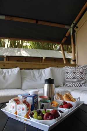 Cozy outdoor breakfast tray on a patio table: white tray with fresh grapes, plums and strawberries, croissants and rolls, sliced cheese, juice cartons, a cereal box, coffee carafe and mugs set in front of a white cushioned sofa under a bamboo canopy.