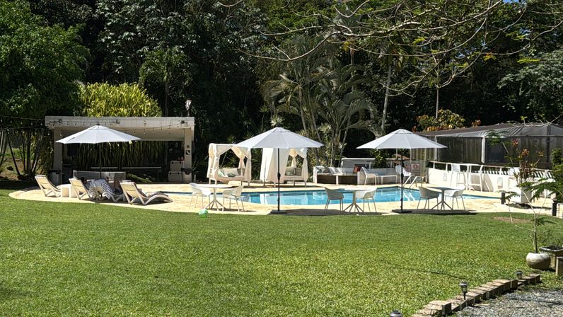 Sunny outdoor pool in a lush tropical garden with white umbrellas, lounge chairs, cabana and manicured green lawn — resort-style relaxation.